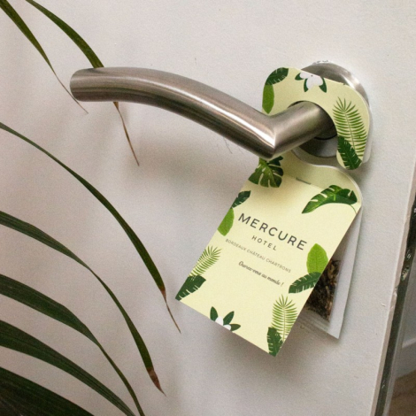 Accroche-porte cartonné vert à motifs végétaux suspendu à une poignée métallique avec marquage visible.