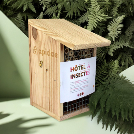 Hôtel à insectes en bois en extérieur avec feuillage en arrière-plan, compartiments en tiges creuses visibles derrière un grilla