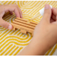 Porte-savon en bois clair personnalisé manipulé par deux mains, posé sur un tissu à motifs avec compte-gouttes au-dessus