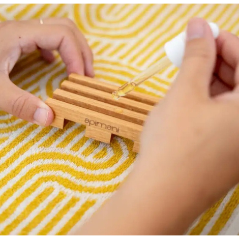Porte-savon en bois clair personnalisé manipulé par deux mains, posé sur un tissu à motifs avec compte-gouttes au-dessus