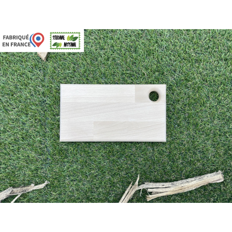 Planche à découper rectangulaire en bois clair, photographiée de face sur fond végétal, avec trou circulaire dans un angle, surf