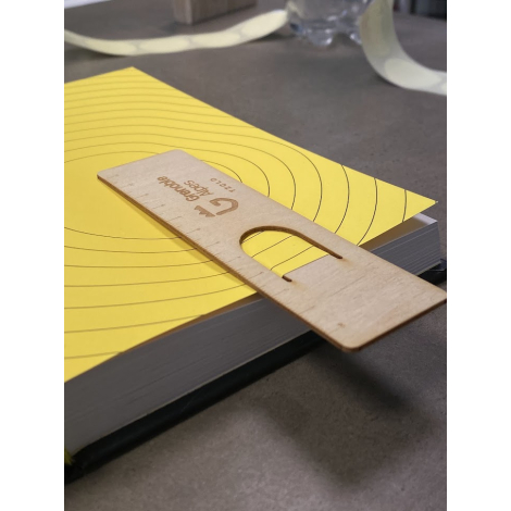 Marque-page en bois inséré dans un livre ouvert, découpe en U engagée sur les pages, gravure visible sur la face supérieure expo