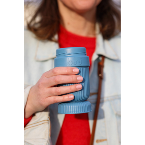 Gros plan sur une personne tenant à la main un mug réutilisable bleu ouvert, à corps rainuré, avec son couvercle placé sous le m