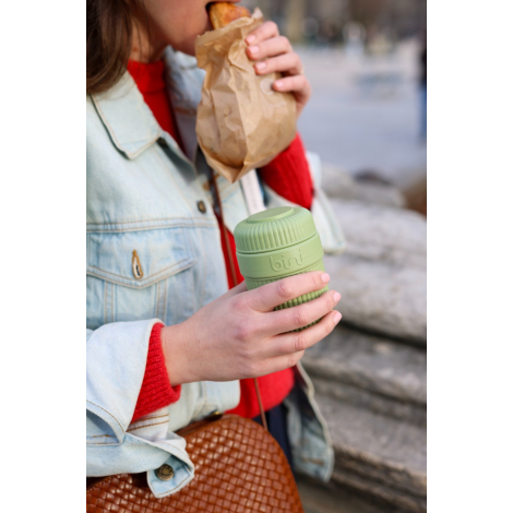 Personne tenant un mug réutilisable vert clair fermé, à corps rainuré et couvercle strié, tout en mangeant une viennoiserie dans