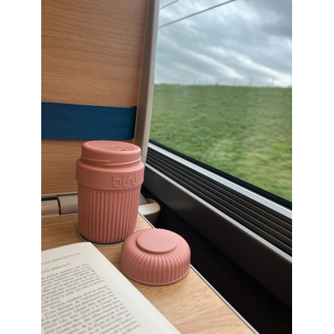 Mug réutilisable rose à corps rainuré posé sur une tablette près de la fenêtre d’un train, avec son couvercle retourné à côté et