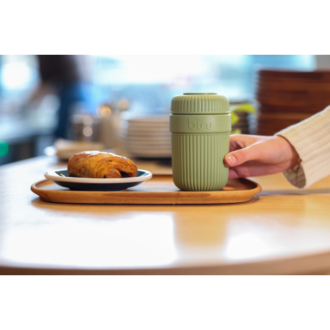 Mug réutilisable vert à corps rainuré et couvercle strié posé sur un plateau à côté d’une viennoiserie, une main placée derrière