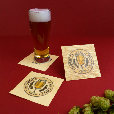 Sous-bocks carrés en bois personnalisés autour d’un verre de bière, présentés avec des fleurs de houblon sur fond rouge.