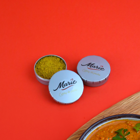 Boîtes rondes métalliques personnalisées contenant des épices, présentées sur fond rouge avec un plat de curry.