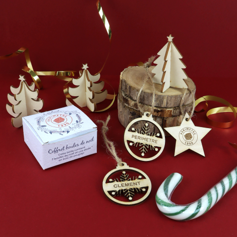 Coffret de Noël et décorations personnalisées en bois mis en scène sur fond rouge avec sapins en bois et rubans dorés.