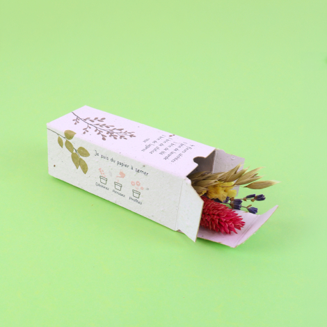 Étui personnalisé en papier à semer contenant un petit bouquet de fleurs séchées, présenté ouvert sur fond vert.